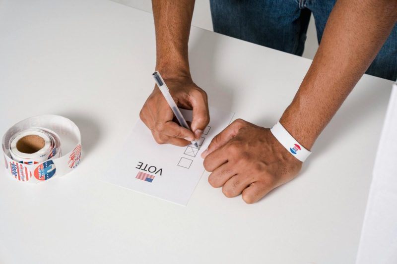 Man's hands filling out a voting ballot with a roll of vote stickers next to his hand.