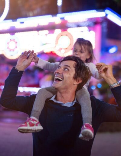 Authentic shot of a happy smiling father carrying his little daughter on a shoulders having fun together in amusement park with luna park lights at night.