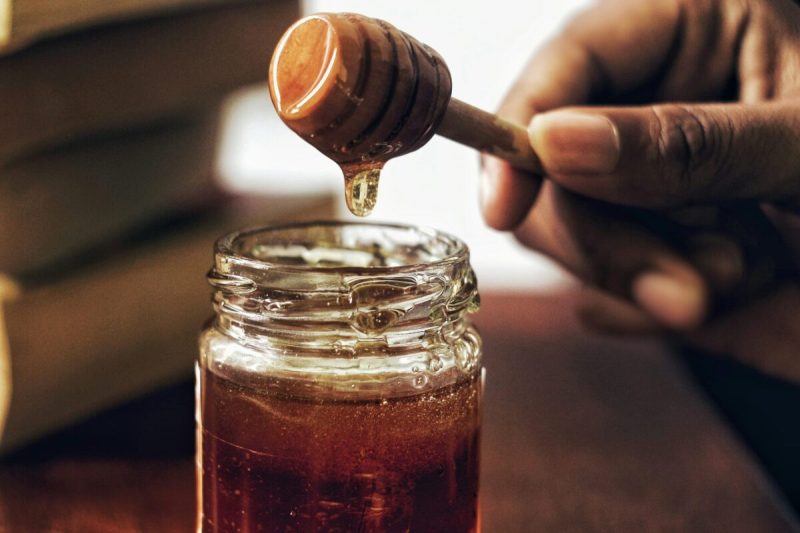 a hand holds a honey dipper outside of a jar of honey