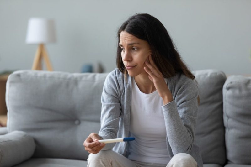 Photo of a woman sitting on the couch, holding a positive pregnancy test and looking disappointed.